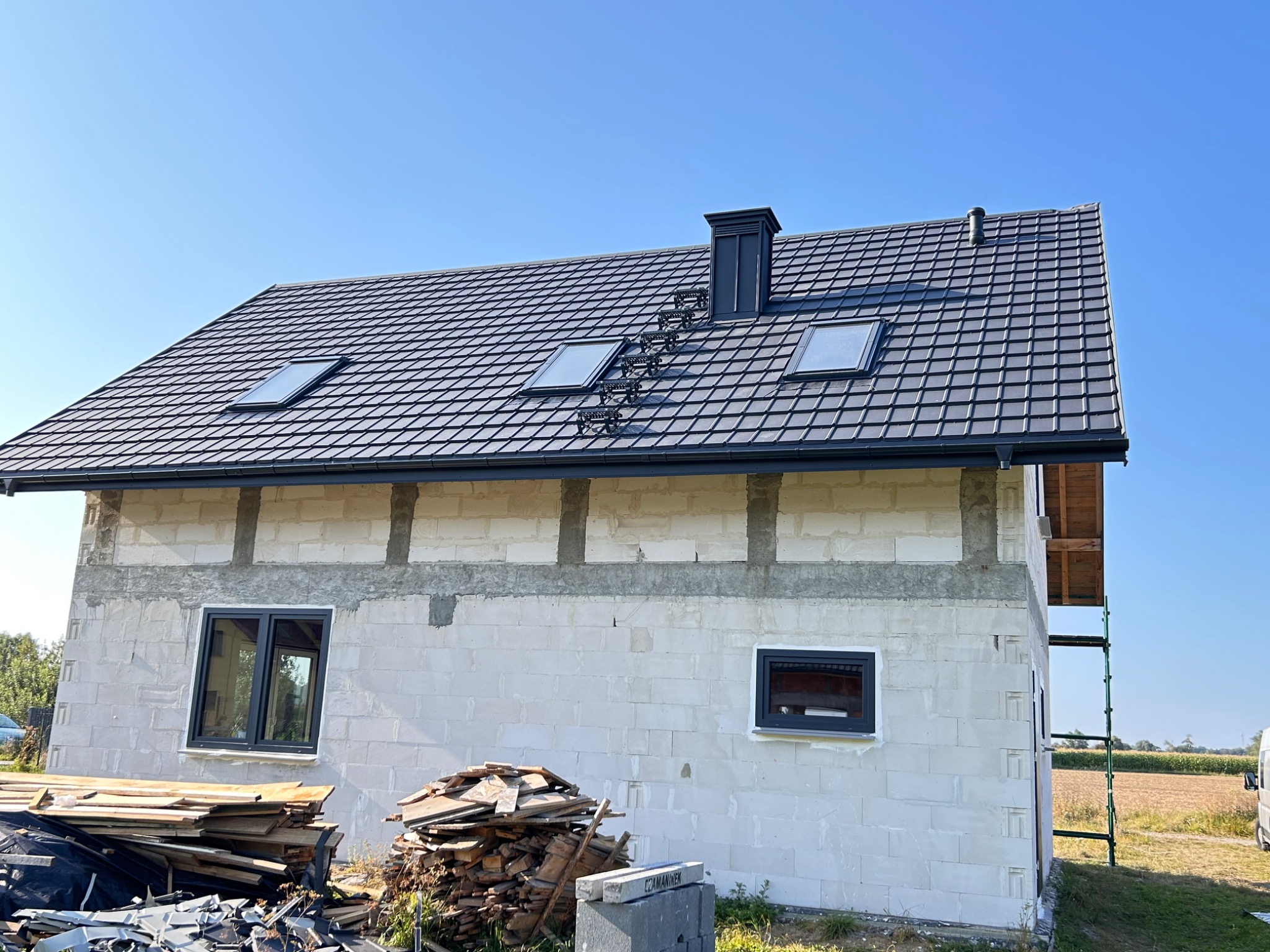 Dom w trakcie budowy z nowym, ciemnoszarym dachem z trzema oknami dachowymi i kominem z drabinką kominiarską, ściany z białego bloczka częściowo otynkowane, widoczne rusztowanie i sterta odpadów...