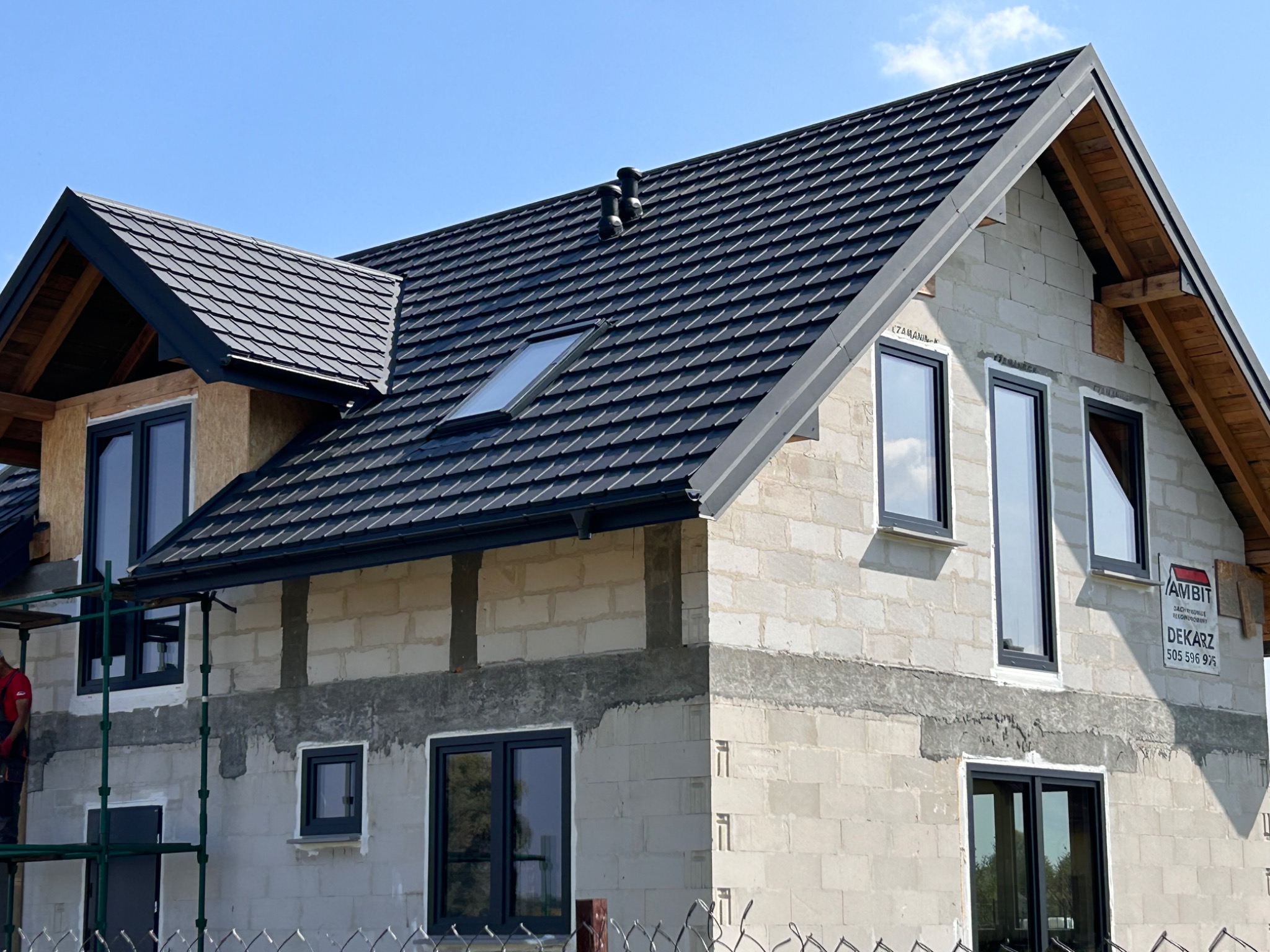 Dom w trakcie budowy z nowym, ciemnoszarym dachem z dachówki modułowej, oknem dachowym i kominkami wentylacyjnymi, widoczny fragment rusztowania i reklama dekarza na ścianie.
