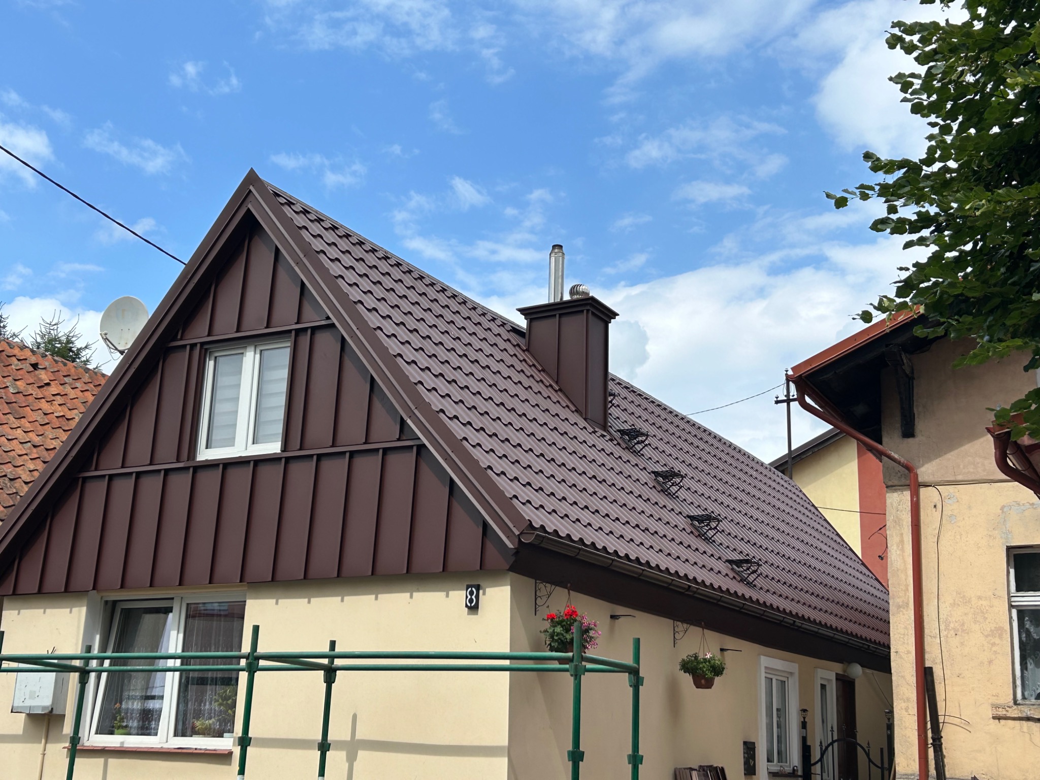 Dom z nowym, brązowym dachem z blachy trapezowej, kominem i rynnami, numer posesji '8', kwiaty w wiszących donicach, częściowo widoczne rusztowanie.