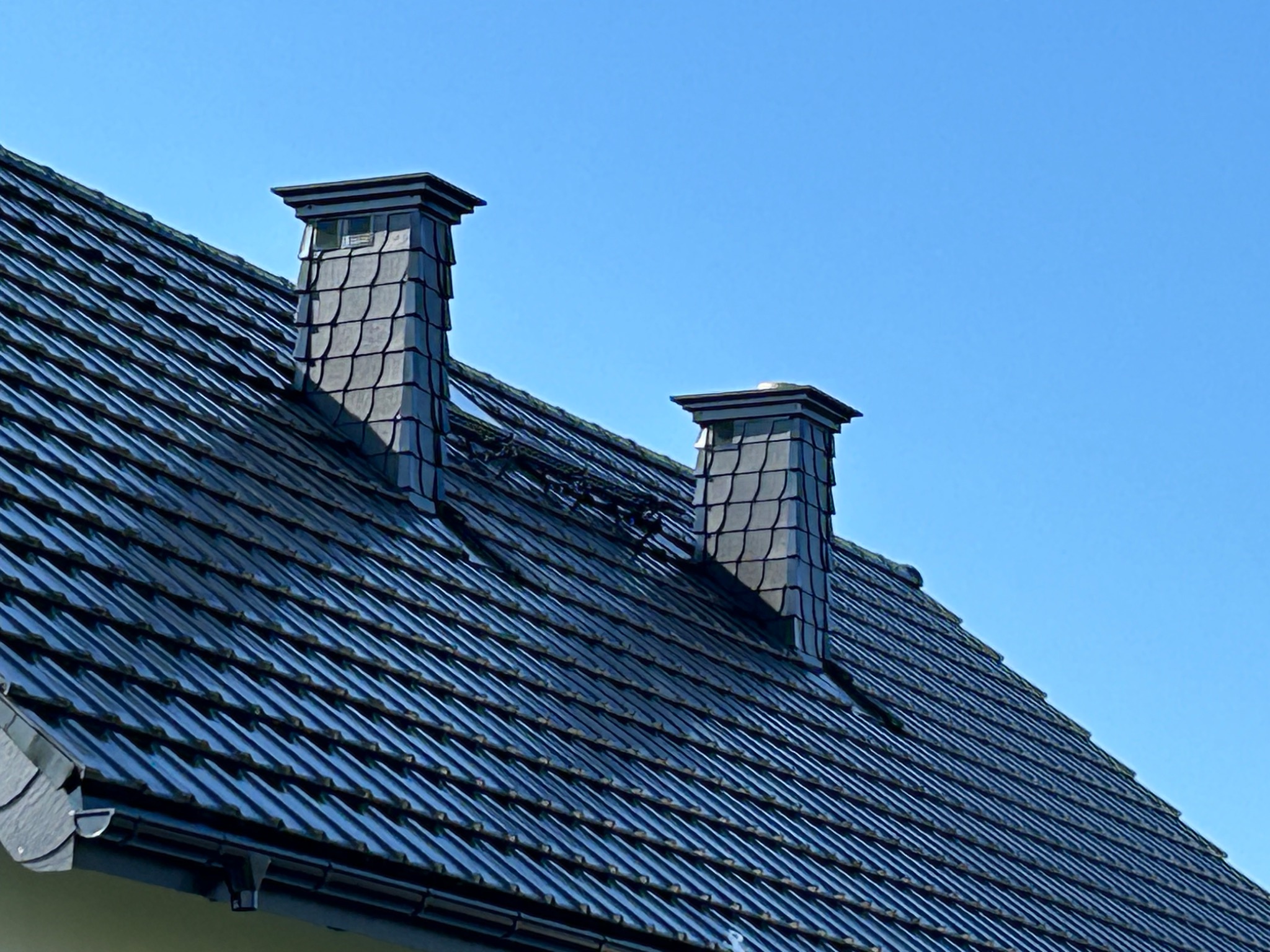 Nowy dach z ciemnej dachówki ceramicznej i dwoma kominami w Gdańsku, widziany pod kątem na tle błękitnego nieba.