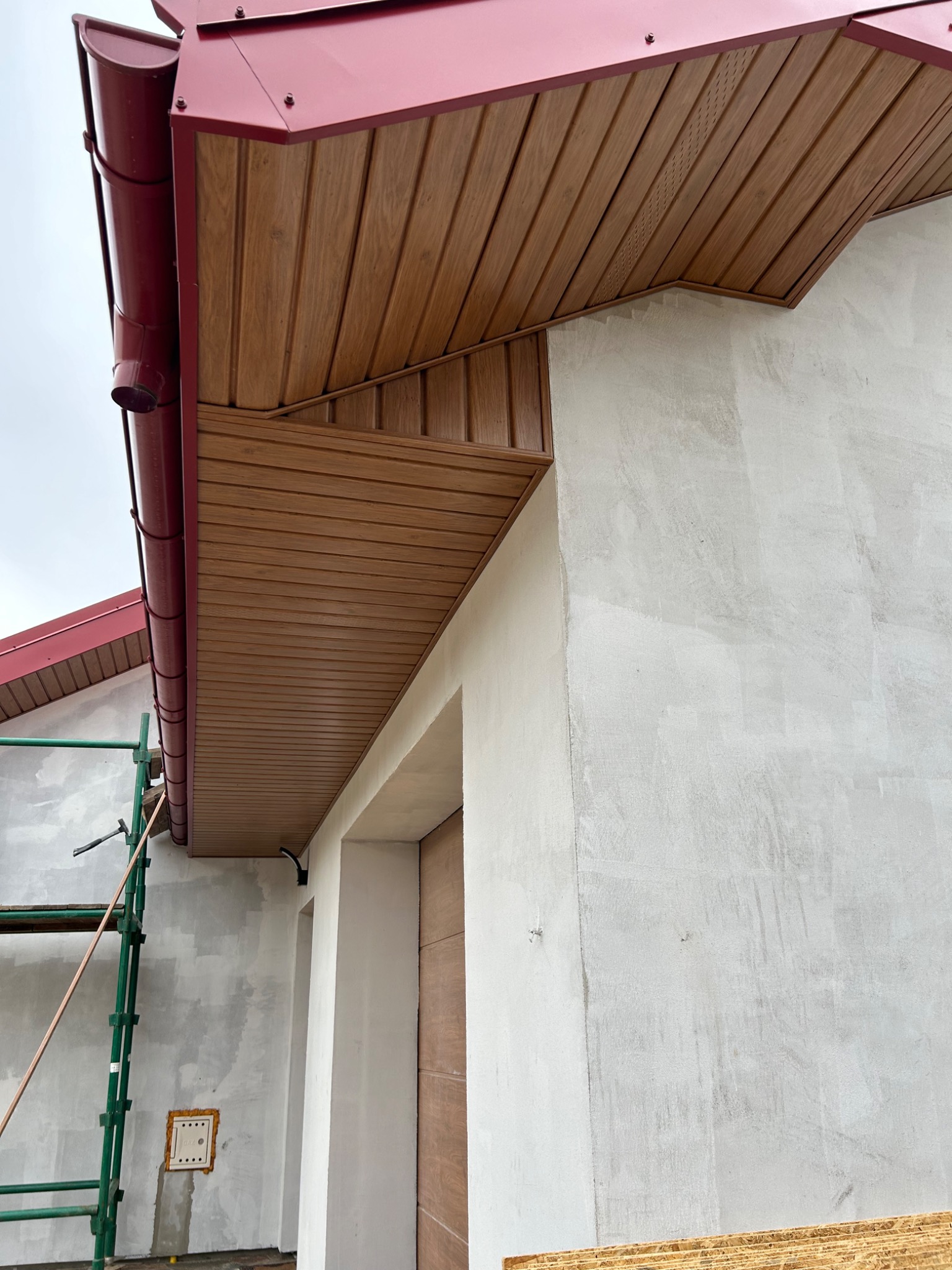 Ciemnoczerwona rynna spustowa i drewnopodobna podbitka dachowa na tle jasnoszarej ściany budynku w trakcie wykończenia, widoczne rusztowanie