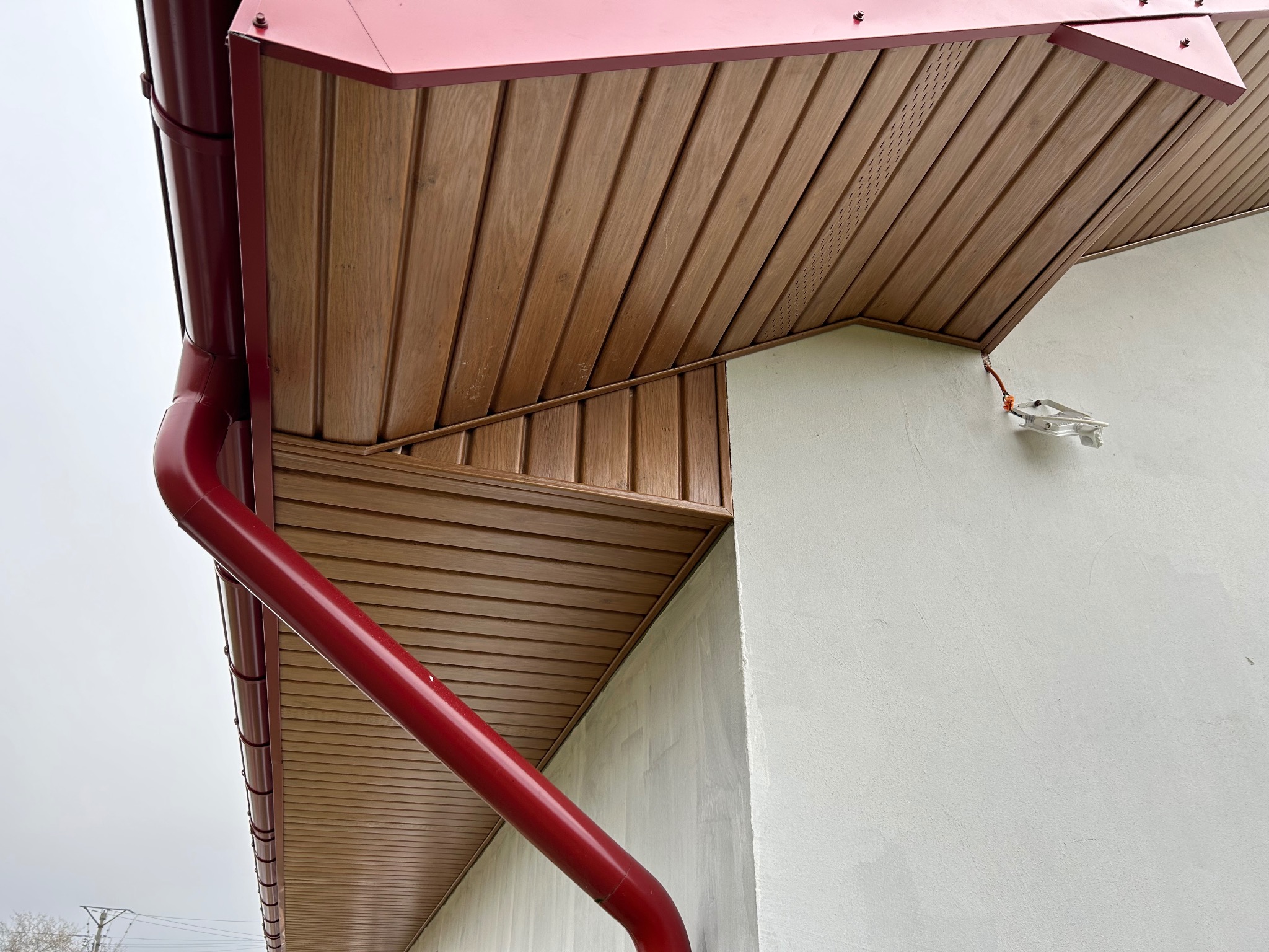 Fragment dachu z drewnianą podbitką, czerwonymi rynnami i elementem oświetlenia zewnętrznego na ścianie budynku.