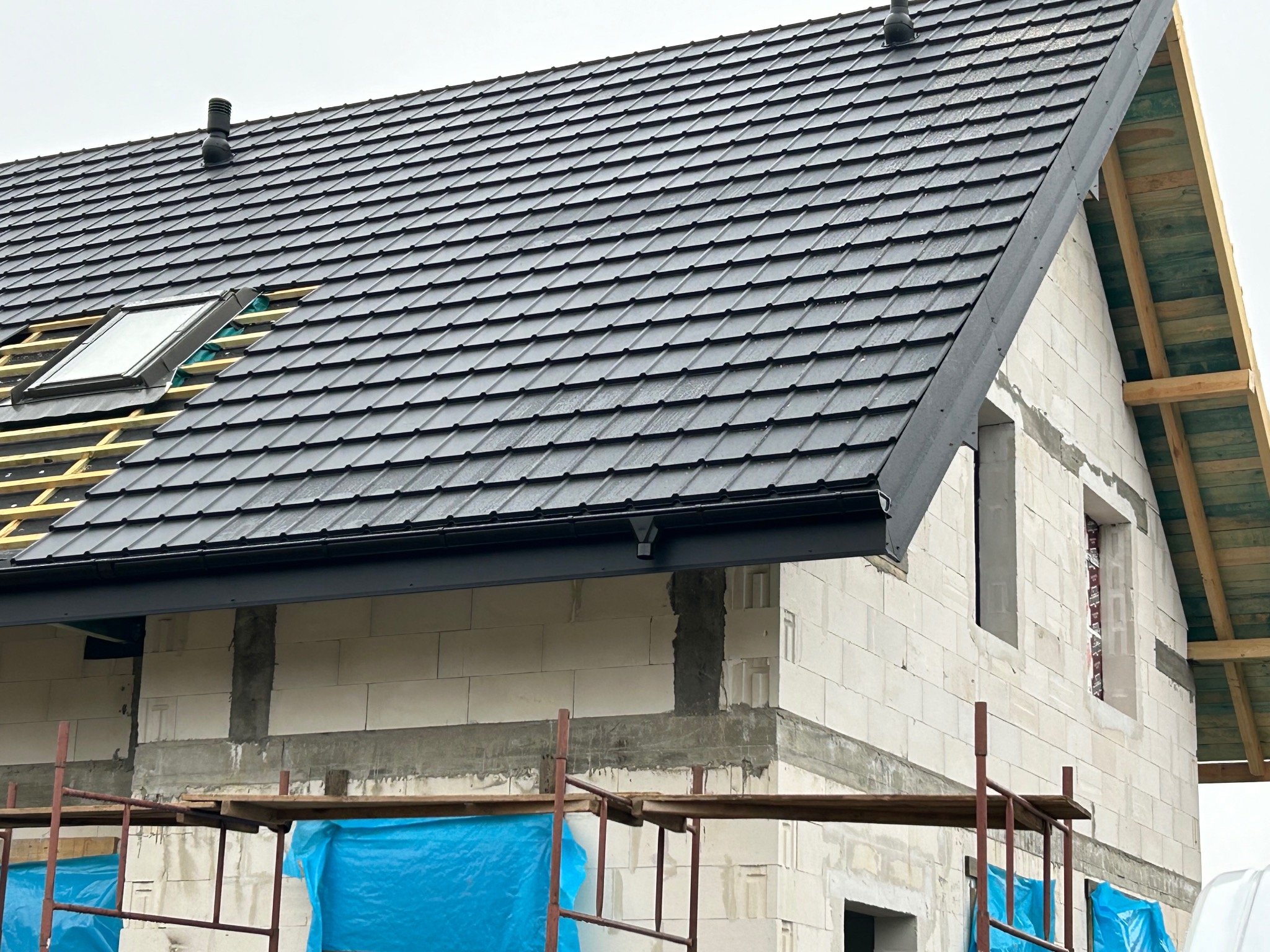 Częściowo pokryty dach z czarnej dachówki ceramicznej na surowym budynku z białego betonu komórkowego, widoczne okno dachowe, rynna, kominek wentylacyjny i rusztowania z niebieską folią...