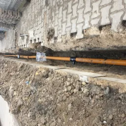 Pomarańczowe rury instalacyjne w wykopie ziemnym wzdłuż ułożonej kostki brukowej, przygotowane do zasypania, widoczne przewody elektryczne wzdłuż rur.