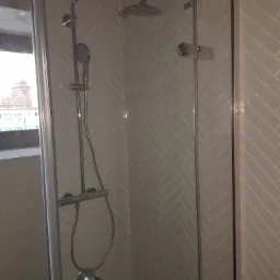 Szklana kabina prysznicowa z chromowaną armaturą na tle ściany wyłożonej jasnymi, ukośnymi płytkami w jodełkę, widok z zewnątrz kabiny, z oknem i budynkiem w tle.