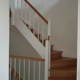 Drewniane schody z białymi balustradami i poręczą z jasnego drewna, widoczne z dołu, po remoncie.