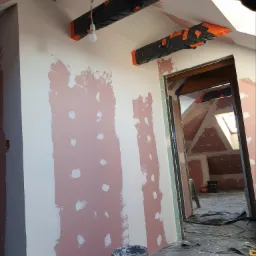 Poddasze w trakcie prac przygotowawczych do wykończenia: ściany z nałożoną warstwą gipsu, widoczne łaty, zabezpieczone folią belki stropowe, podłoga zakryta folią ochronną, wiadro z masą szpachlową...