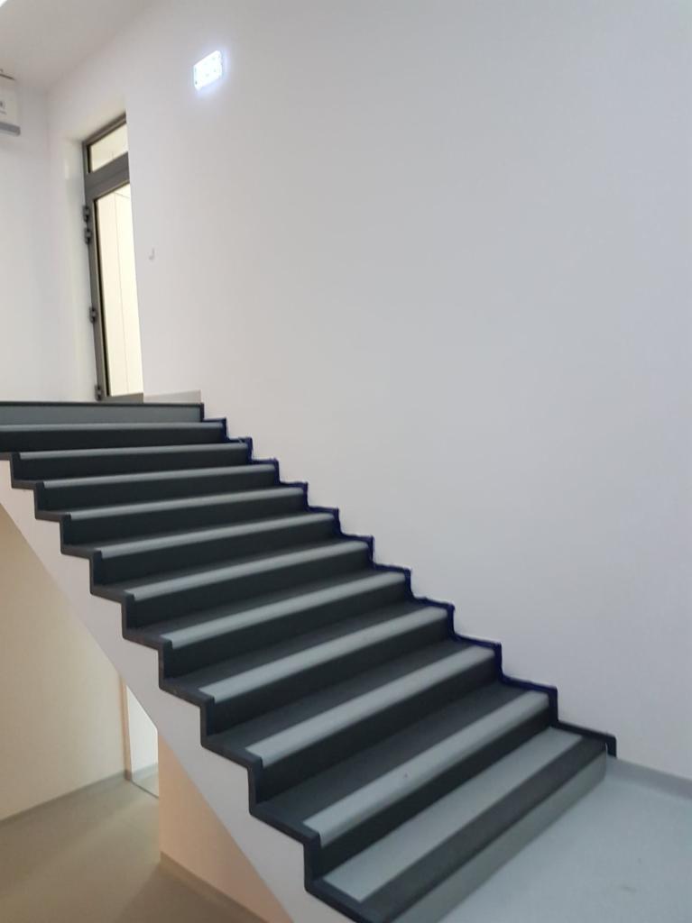 Schody wewnętrzne z szarymi stopniami i białymi podstopnicami, prowadzące w górę, widoczne drzwi i biała ściana.