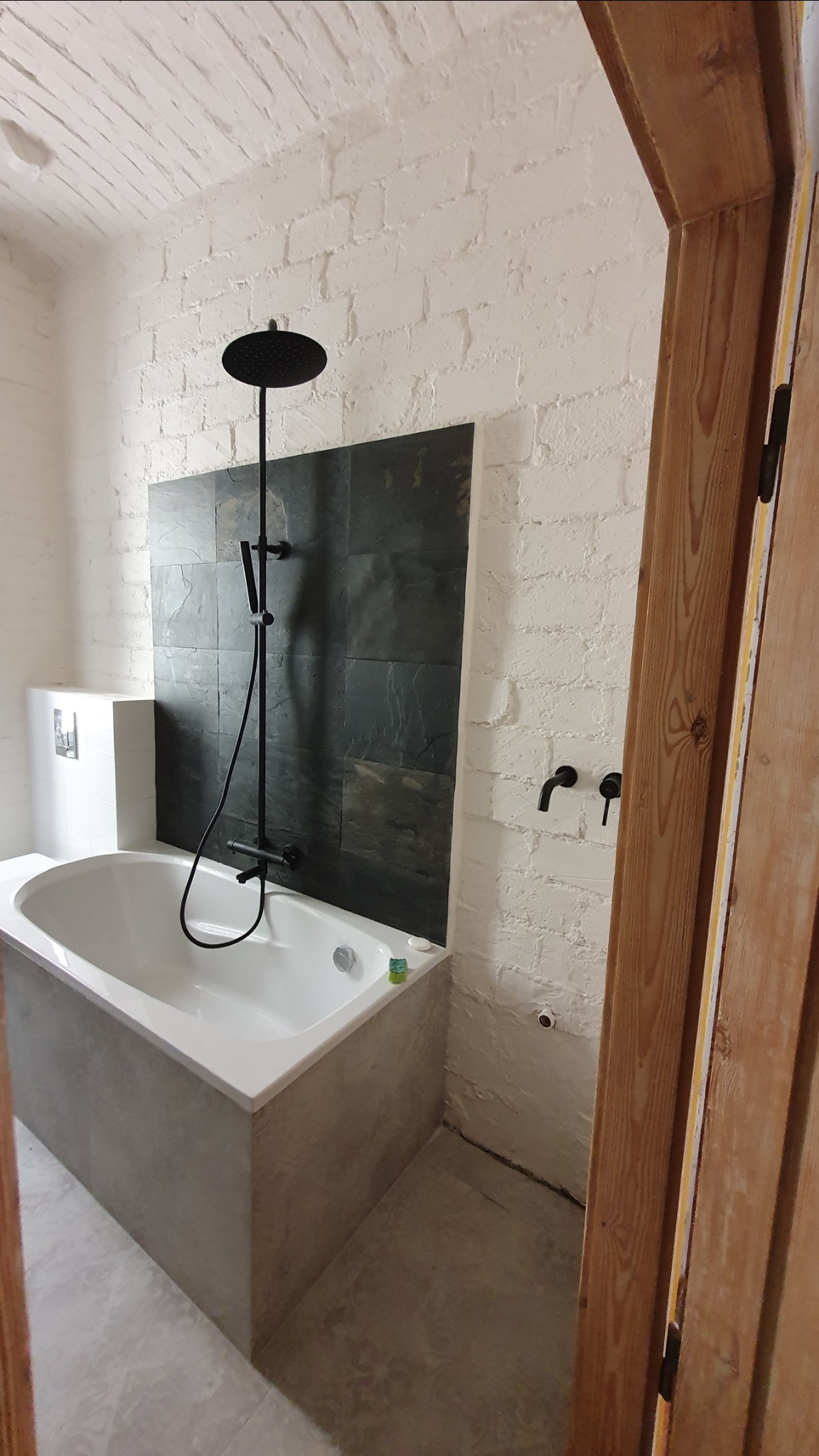 Nowoczesna łazienka z białą cegłą, czarną armaturą i wanną obłożoną szarą płytą imitującą beton, widok zza drewnianych drzwi.