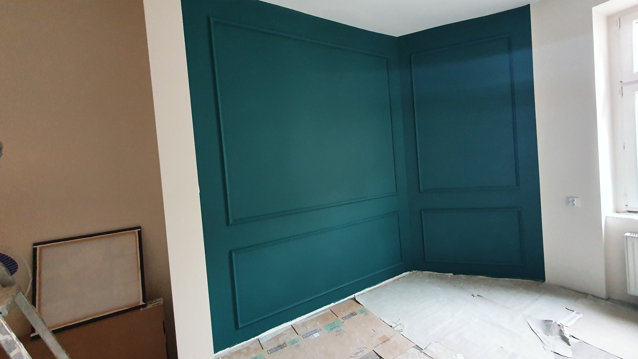 Ściana pomalowana na ciemnozielony kolor z dekoracyjnymi listwami, podłoga zabezpieczona kartonem, widoczny fragment grzejnika i okna.