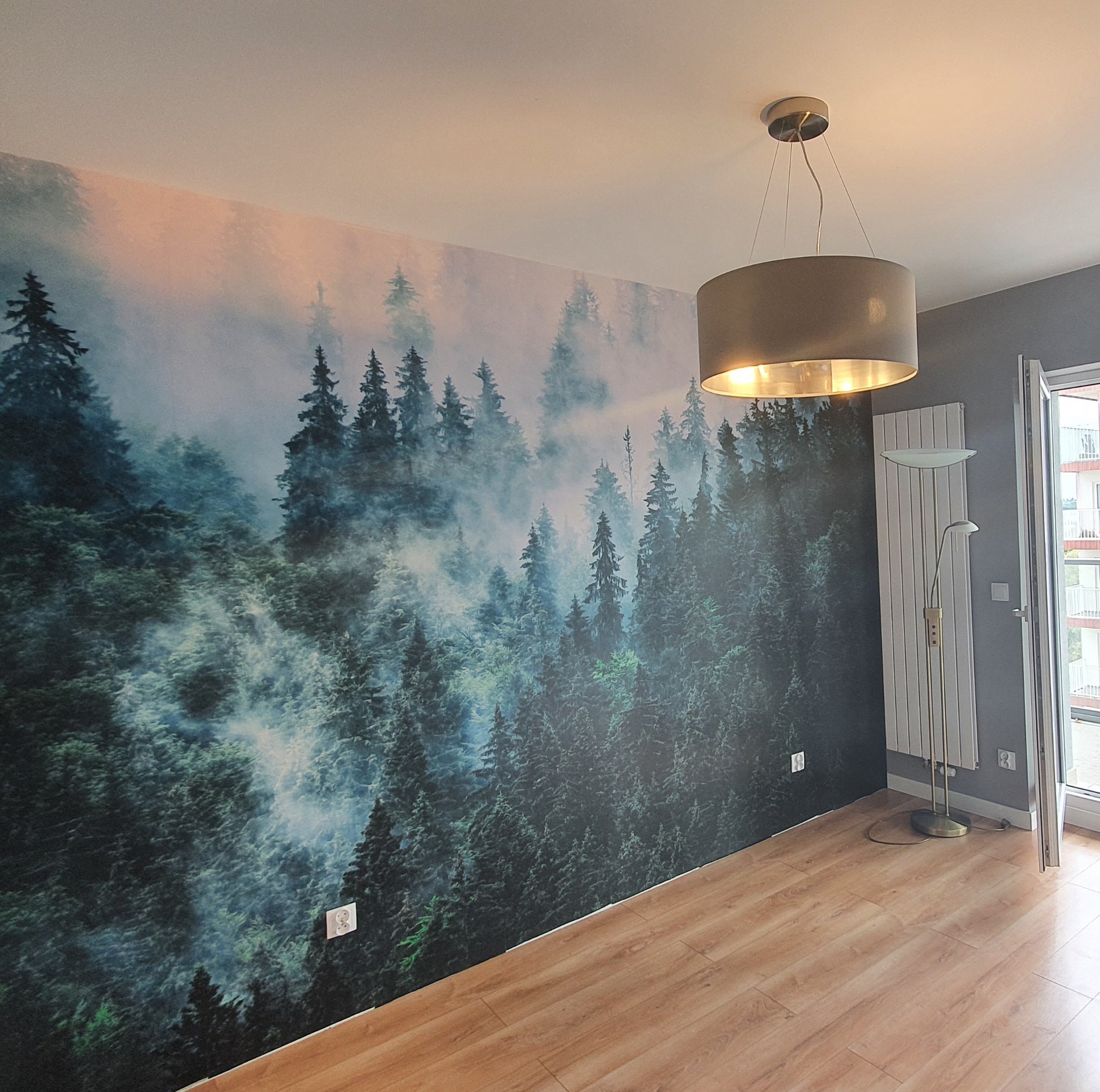 Pokój z fototapetą przedstawiającą mglisty las iglasty, oświetlony wiszącą lampą z szarym abażurem i uzupełniony podłogą z paneli w odcieniach drewna.
