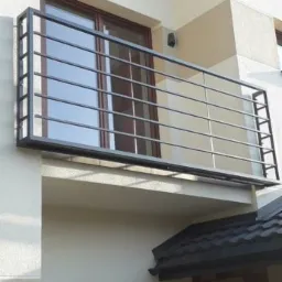 Prosta, metalowa balustrada balkonowa z poziomymi prętami zamontowana na jasnej elewacji budynku z widocznym oknem i fragmentem dachu.