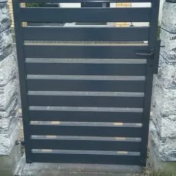 Nowoczesna, grafitowa furtka z poziomymi panelami, zamontowana między kamiennymi słupkami, widok z bliska na trawnik przed wejściem.