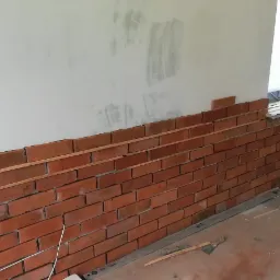 Częściowo wykończona ściana z cegły klinkierowej wewnątrz pomieszczenia, widoczne drewniane listwy poziomujące i narzędzia glazurnicze na podłodze, surowy stan ściany w górnej części.
