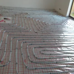 Szare rury ogrzewania podłogowego ułożone spiralnie na czerwonej siatce z folii, widok z góry, w tle ściana i okno.