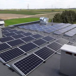 Rzędy paneli słonecznych na płaskim dachu budynku, komin wentylacyjny i odległy krajobraz z liniami wysokiego napięcia w tle.