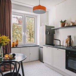 Monika Wierzba-Krygiel Architektura i Wnętrza - Jasna kuchnia z czarnymi akcentami: okrągły stół z bukietem żółtych kwiatów, białe szafki z czarnym blatem, czarna armatura i piekarnik, pomarańczowa lampa, widok z okna na drzewa.