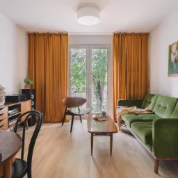 Salon w stylu retro z zieloną sofą, drewnianym stolikiem kawowym, gramofonem i kolumnami głośnikowymi, oświetlony naturalnym światłem wpadającym przez okno z pomarańczowymi zasłonami.