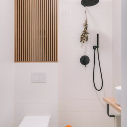 Monika Wierzba-Krygiel Architektura i Wnętrza - Nowoczesna łazienka z białymi płytkami, drewnianą zabudową nad toaletą, czarną armaturą prysznicową i dekoracyjną rzeźbą pływaka w pomarańczowym czepku.