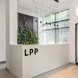 Biała lada recepcyjna z logo LPP, ozdobiona pnącą roślinnością na tle czarnej, perforowanej ściany, oświetlona wiszącymi lampami geometrycznymi i światłem dziennym zza żaluzji.