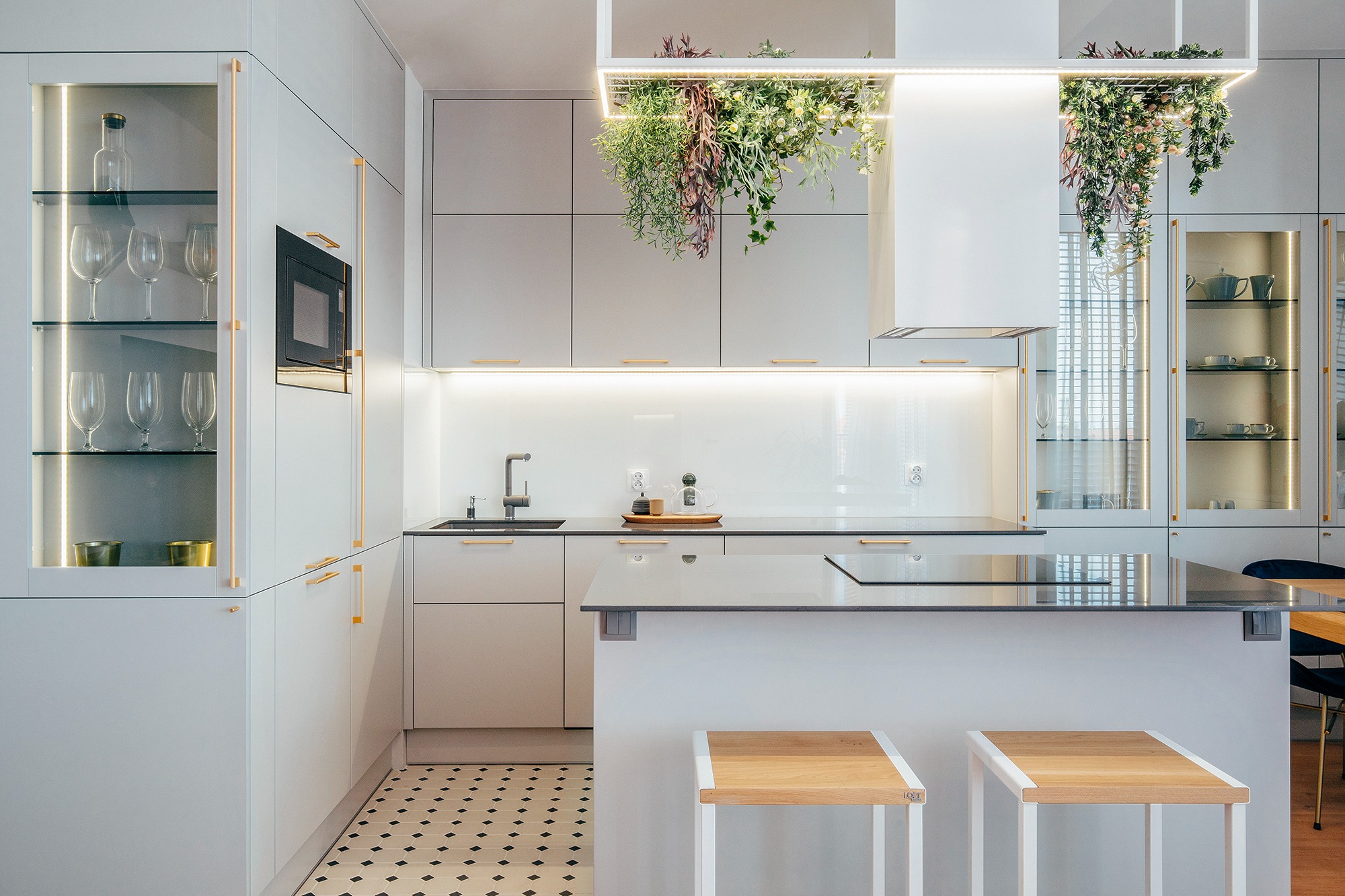 Nowoczesna, jasna kuchnia w Gdańsku z wyspą i dekoracyjnymi roślinami zwisającymi z sufitu, oświetlona punktowo, z czarno-białą mozaiką na podłodze.
