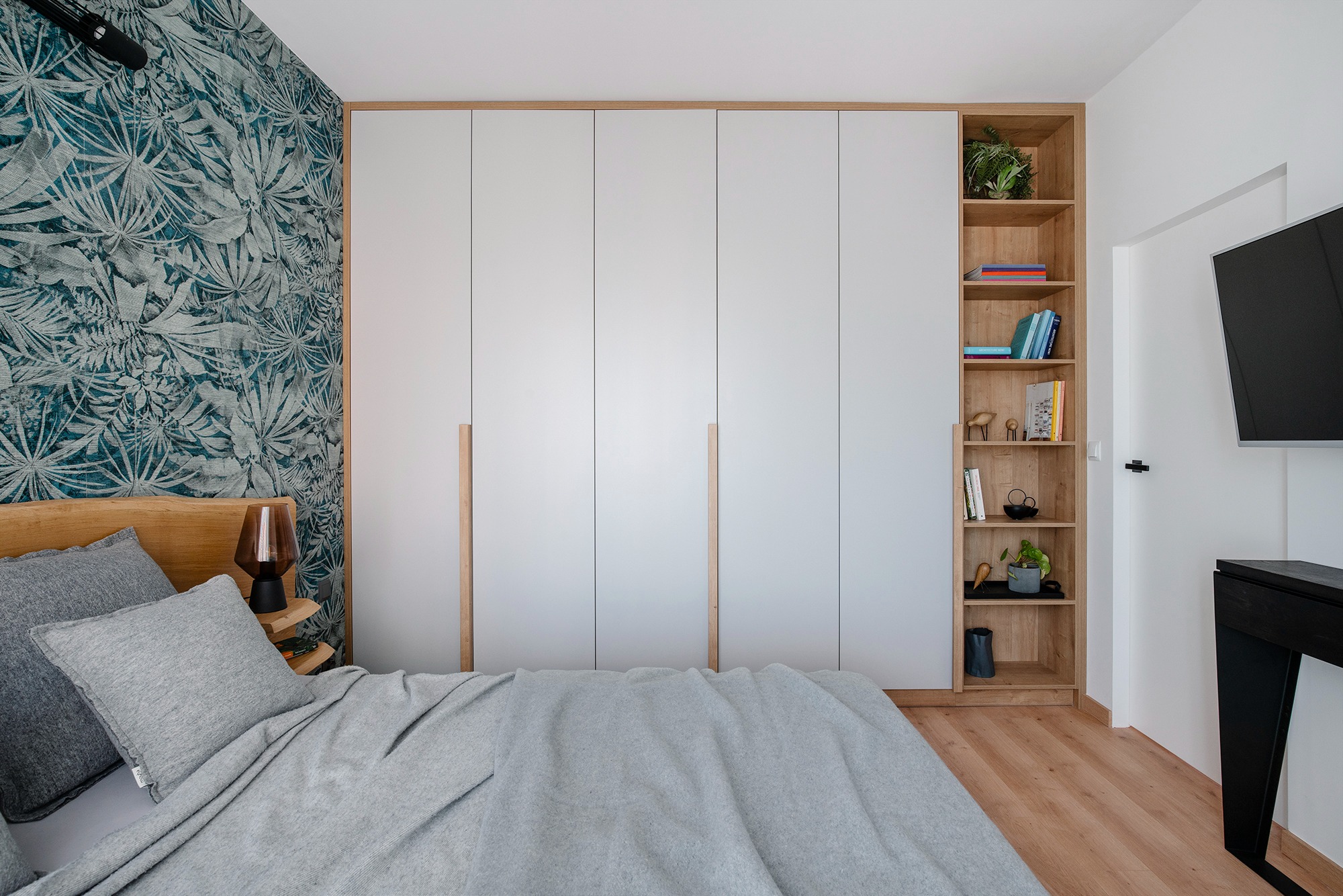 Sypialnia z łóżkiem, szarą pościelą, szafą w zabudowie z drewnianymi uchwytami, regałem z książkami i roślinami, oraz ścianą z tapetą w motywy roślinne.