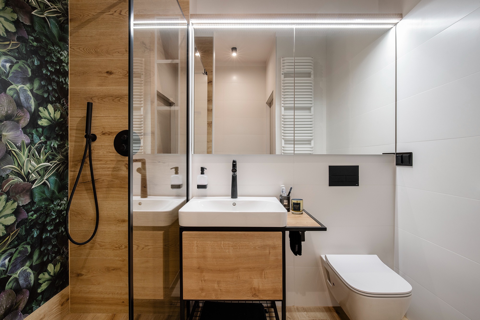 Nowoczesna łazienka z drewnianymi akcentami, kwadratową umywalką nablatową, czarną armaturą, toaletą podwieszaną i ścianą z motywem roślinnym.