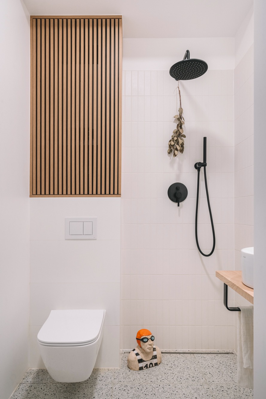 Nowoczesna łazienka z białymi płytkami, drewnianą zabudową nad toaletą, czarną armaturą prysznicową i dekoracyjną rzeźbą pływaka w pomarańczowym czepku.