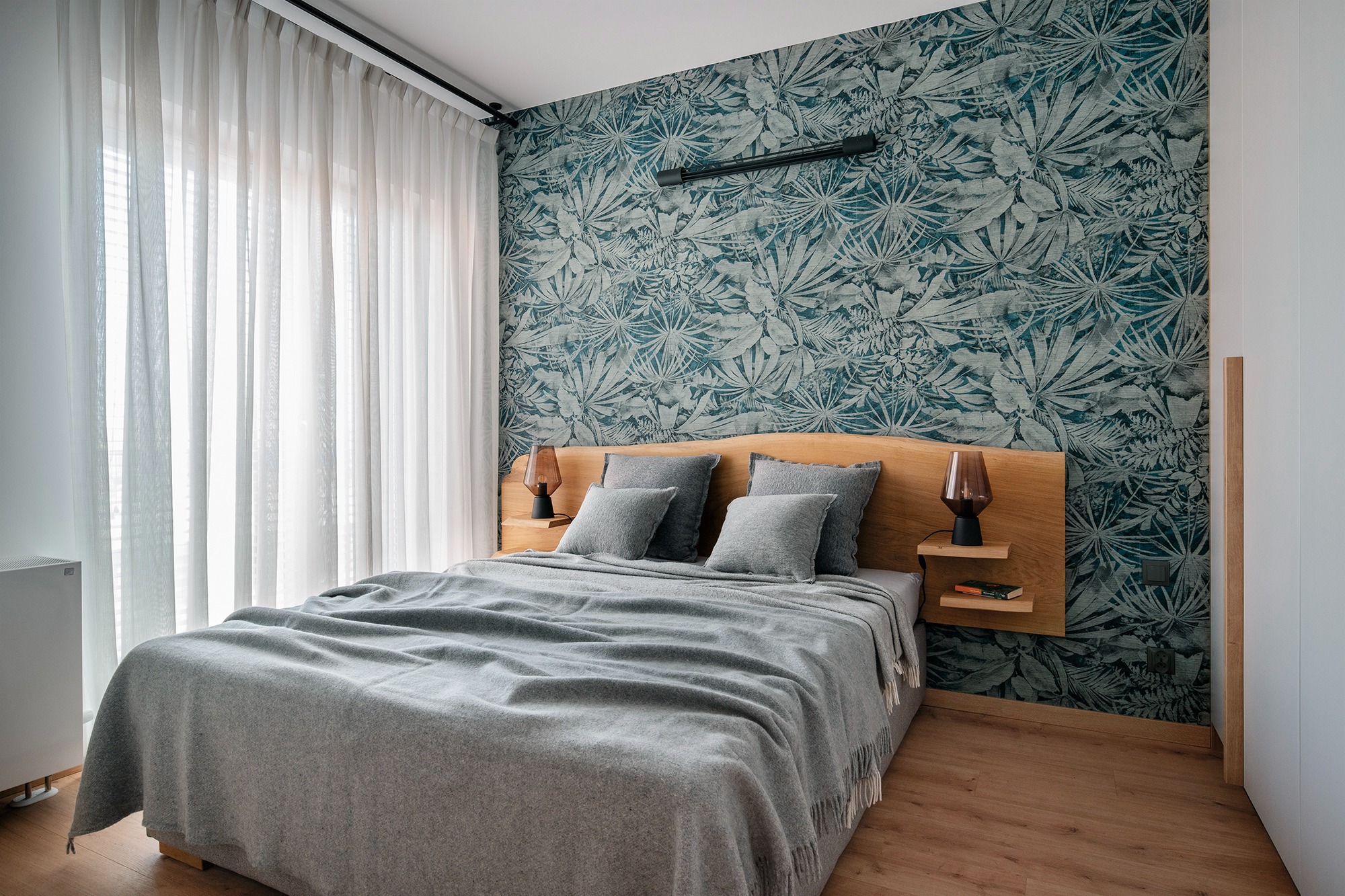 Sypialnia z drewnianym wezgłowiem, szarą narzutą i poduszkami, fototapeta w tropikalne liście oraz lampki nocne z brązowymi abażurami.