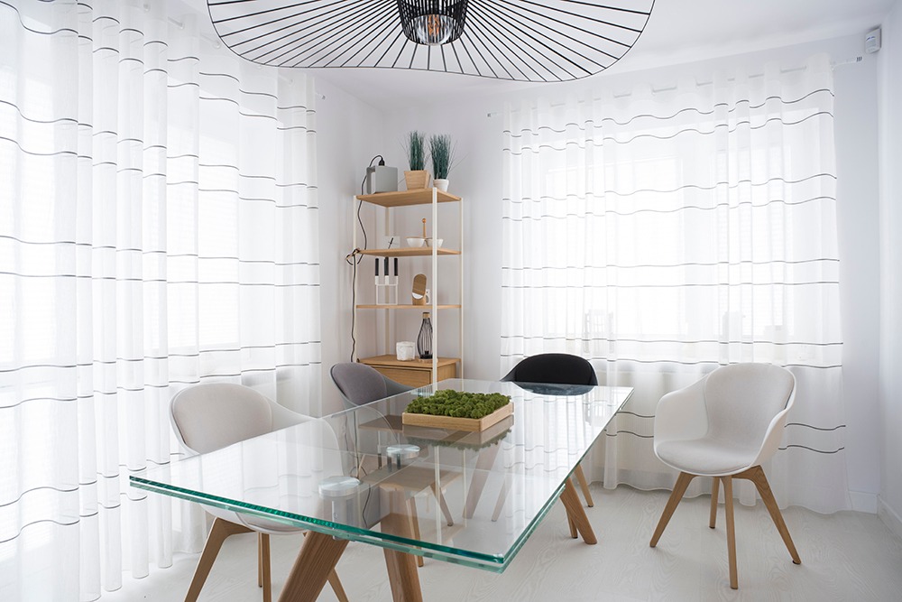 Jasne wnętrze jadalni z szklanym stołem, drewnianymi nogami krzeseł i półką z dekoracjami, oświetlone przez designerską lampę sufitową i okna z delikatnymi, wzorzystymi firanami.