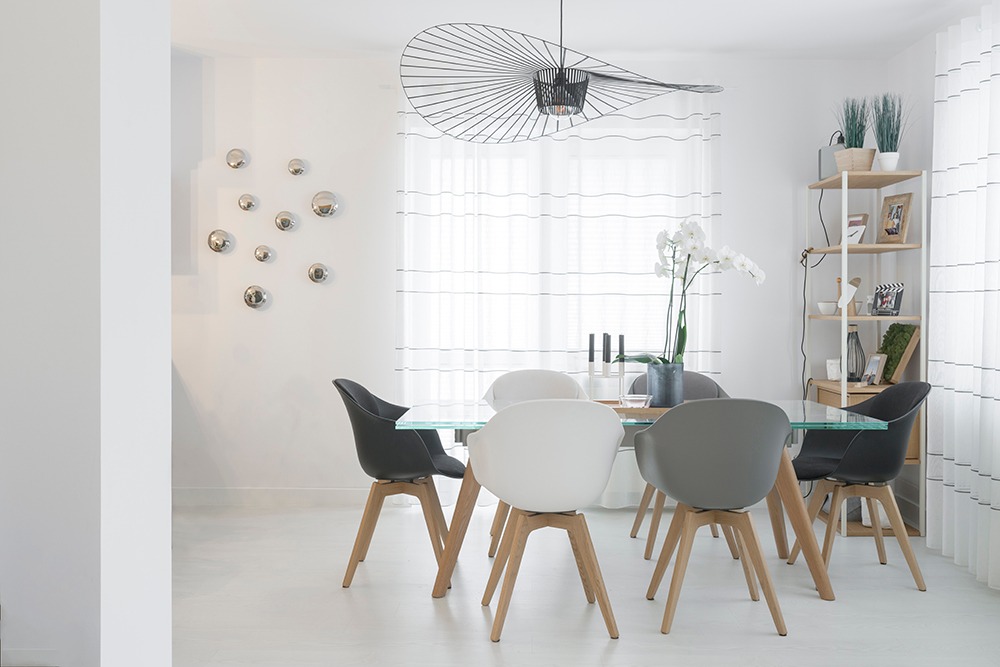 Jasna jadalnia z szklanym stołem otoczonym krzesłami w odcieniach szarości i bieli, ozdobiona wiszącą lampą o ażurowym kształcie i srebrnymi kulami na ścianie.