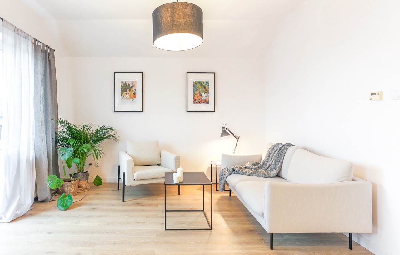 Jasne wnętrze salonu z beżową sofą, fotelem, stolikiem kawowym z czarnym blatem, roślinami doniczkowymi, lampą i grafikami na ścianie.