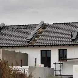 Dom w trakcie budowy z częściowo położonym ciemnoszarym dachem z dachówki ceramicznej, widoczne fragmenty więźby dachowej i rusztowania przy ścianie budynku.