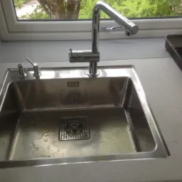 Błyszczący, stalowy zlew kuchenny z chromowaną baterią i dozownikiem mydła, zamontowany przy oknie z widokiem na zieleń, obok suszarka do naczyń.