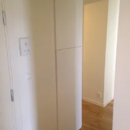 Korytarz z drewnianą podłogą, białe ściany, domofon przy drzwiach wejściowych i widoczne gniazdko elektryczne w ścianie.