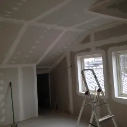 Poddasze w trakcie prac wykończeniowych: płyty gipsowo-kartonowe zamontowane na suficie i ścianach, widoczne łączenia i szpachlowane wkręty, aluminiowa drabina oparta o ścianę pod oknem, w tle...