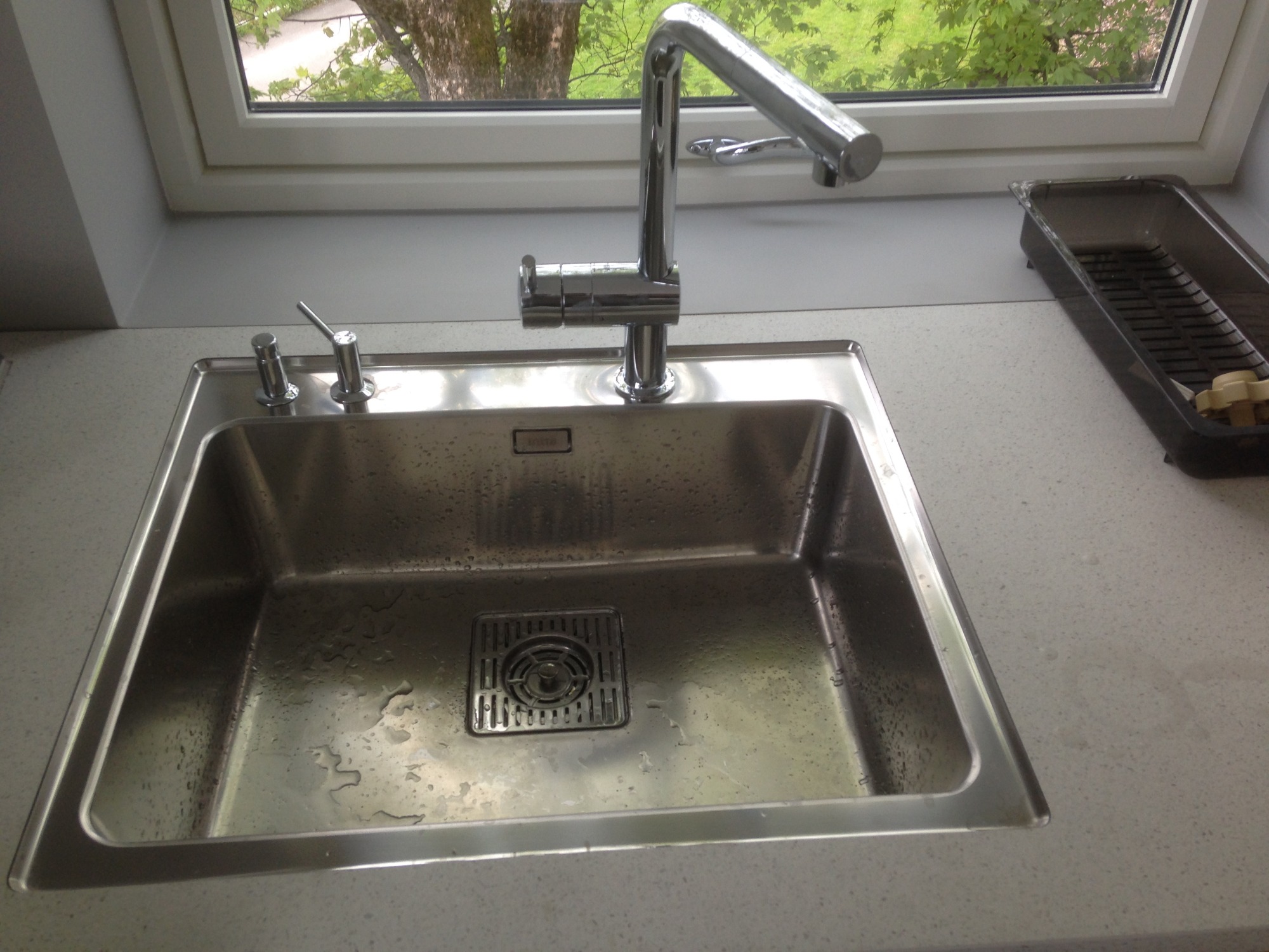 Błyszczący, stalowy zlew kuchenny z chromowaną baterią i dozownikiem mydła, zamontowany przy oknie z widokiem na zieleń, obok suszarka do naczyń.