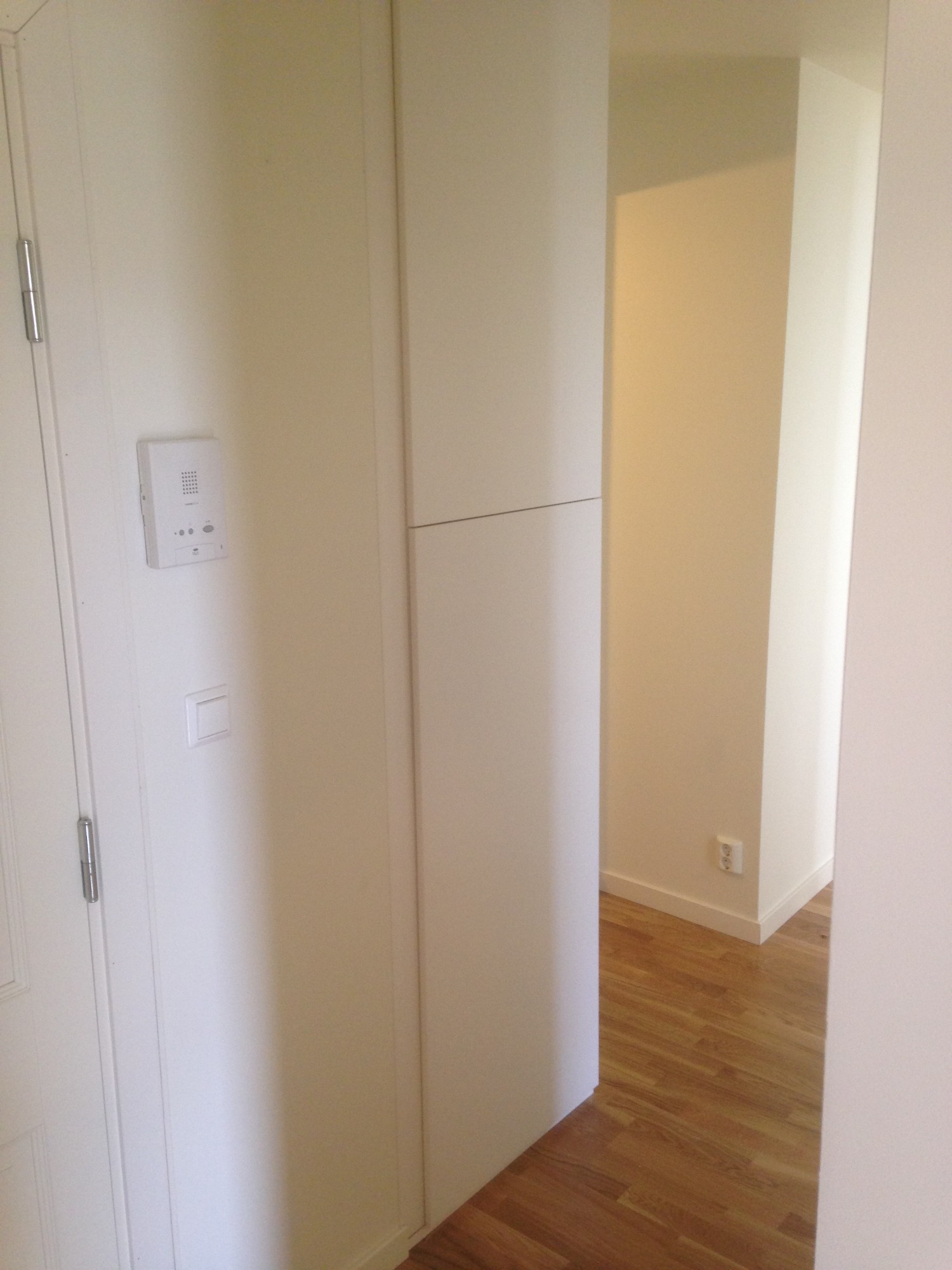 Korytarz z drewnianą podłogą, białe ściany, domofon przy drzwiach wejściowych i widoczne gniazdko elektryczne w ścianie.