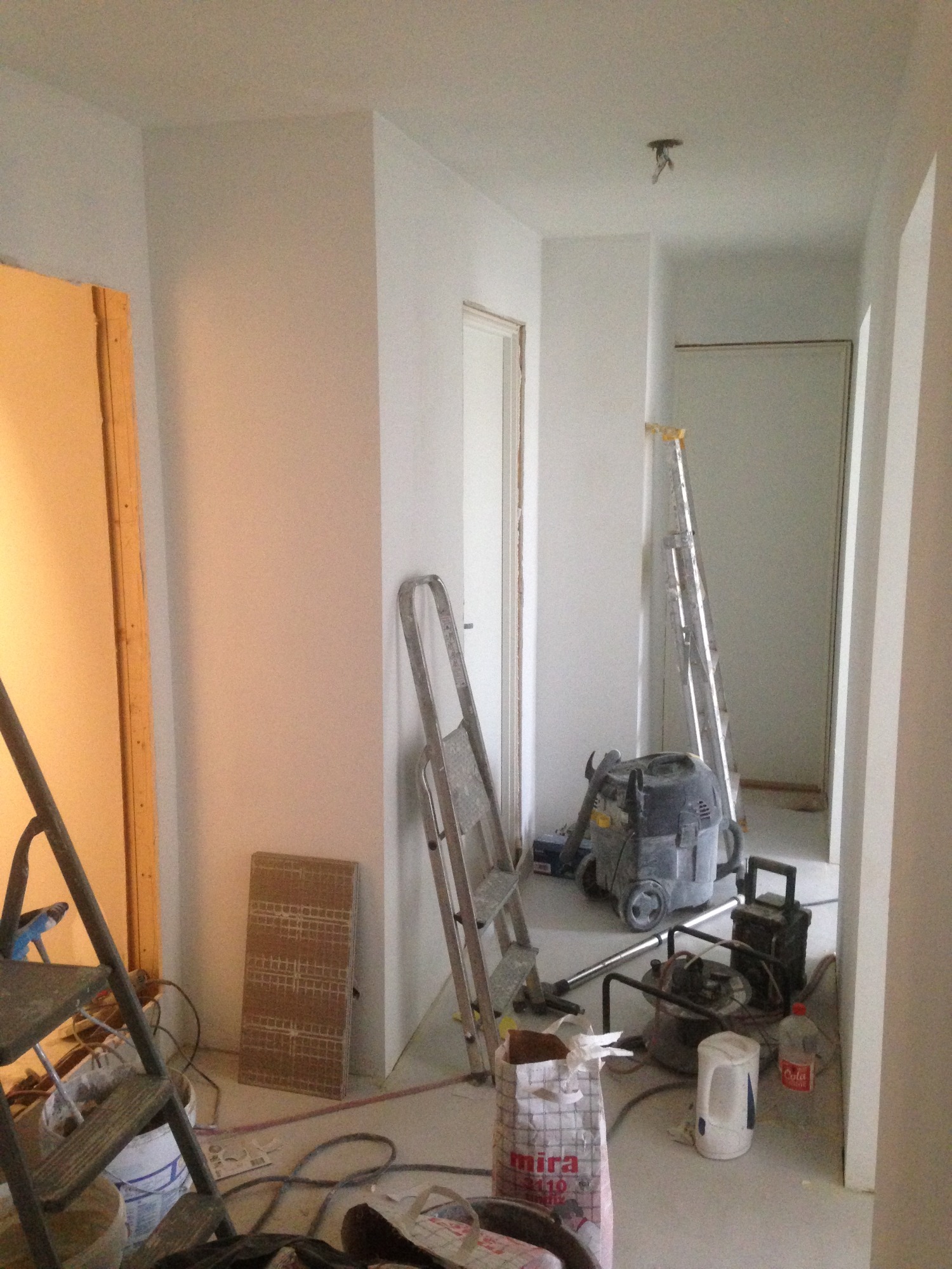 Pokój w trakcie remontu: świeżo wyszpachlowane ściany, drabiny, odkurzacz przemysłowy, wiadro z zaprawą, kabel na podłodze, widoczne ślady po pracach wykończeniowych.