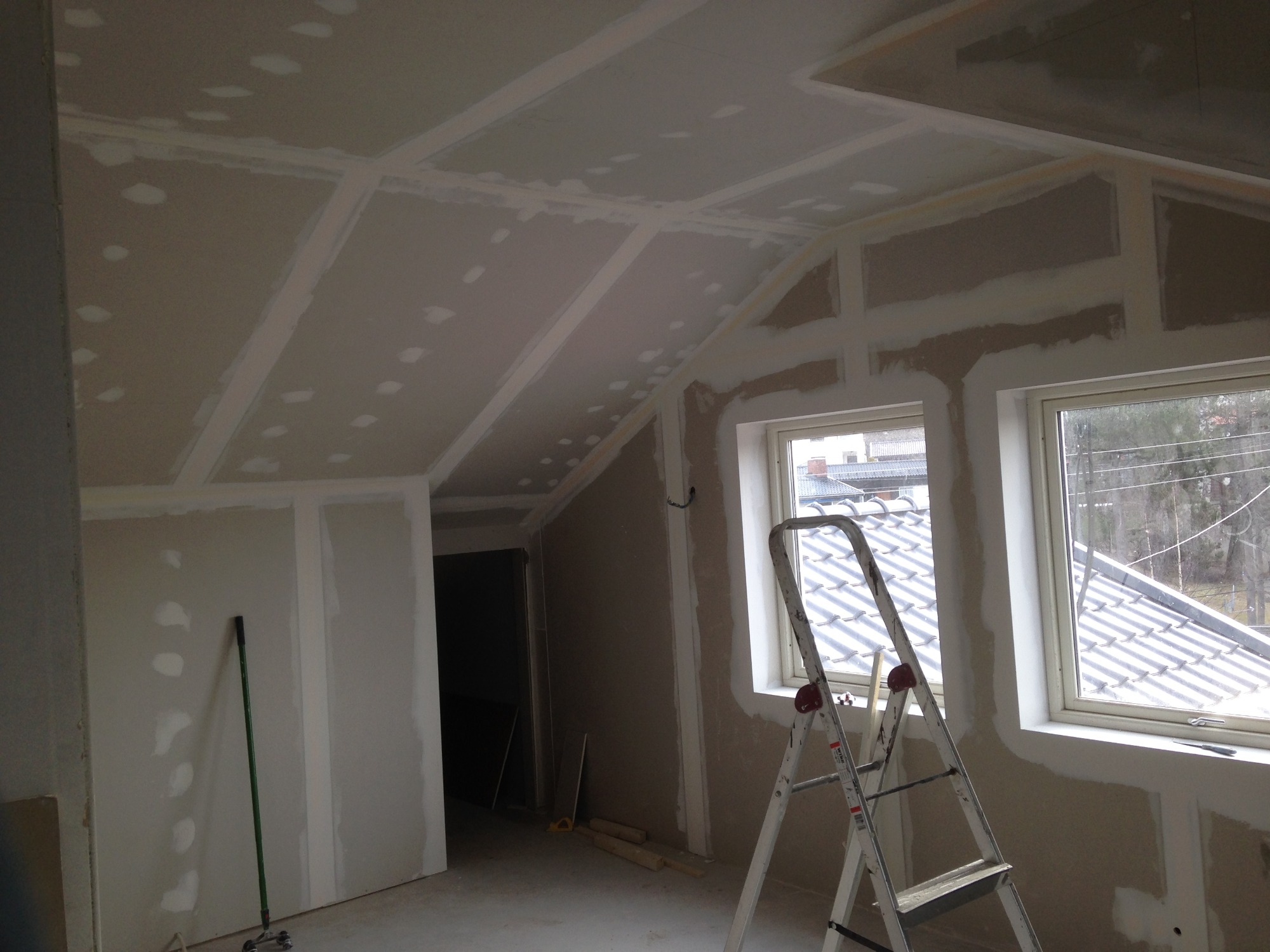 Poddasze w trakcie prac wykończeniowych: płyty gipsowo-kartonowe zamontowane na suficie i ścianach, widoczne łączenia i szpachlowane wkręty, aluminiowa drabina oparta o ścianę pod oknem, w tle...