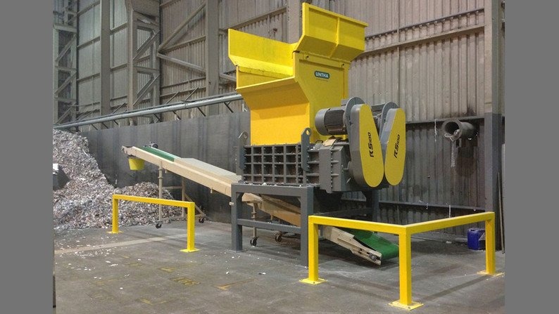 Żółto-szara przemysłowa niszczarka UNTHA RS100 z podajnikiem taśmowym i stertą pociętych materiałów w tle hali.