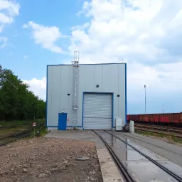 Nowoczesny budynek hali przemysłowej z bramą rolowaną i drabiną na zewnątrz, usytuowany na terenie kolejowym z widocznymi wagonami towarowymi w tle.