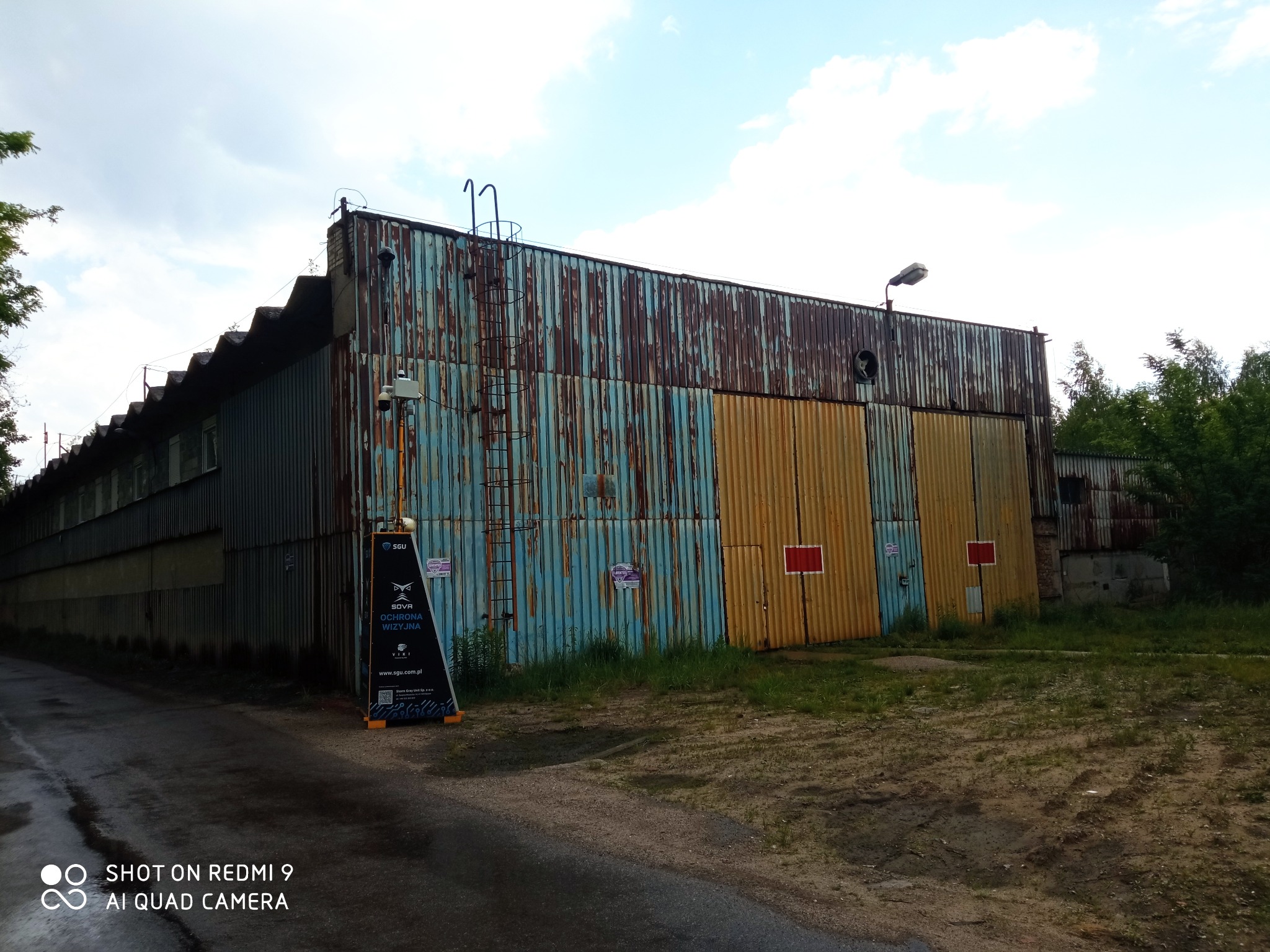 Długi, blaszany budynek magazynowy z widoczną korozją, drabina na elewacji, kamera przemysłowa i tablica informacyjna ochrony wizyjnej