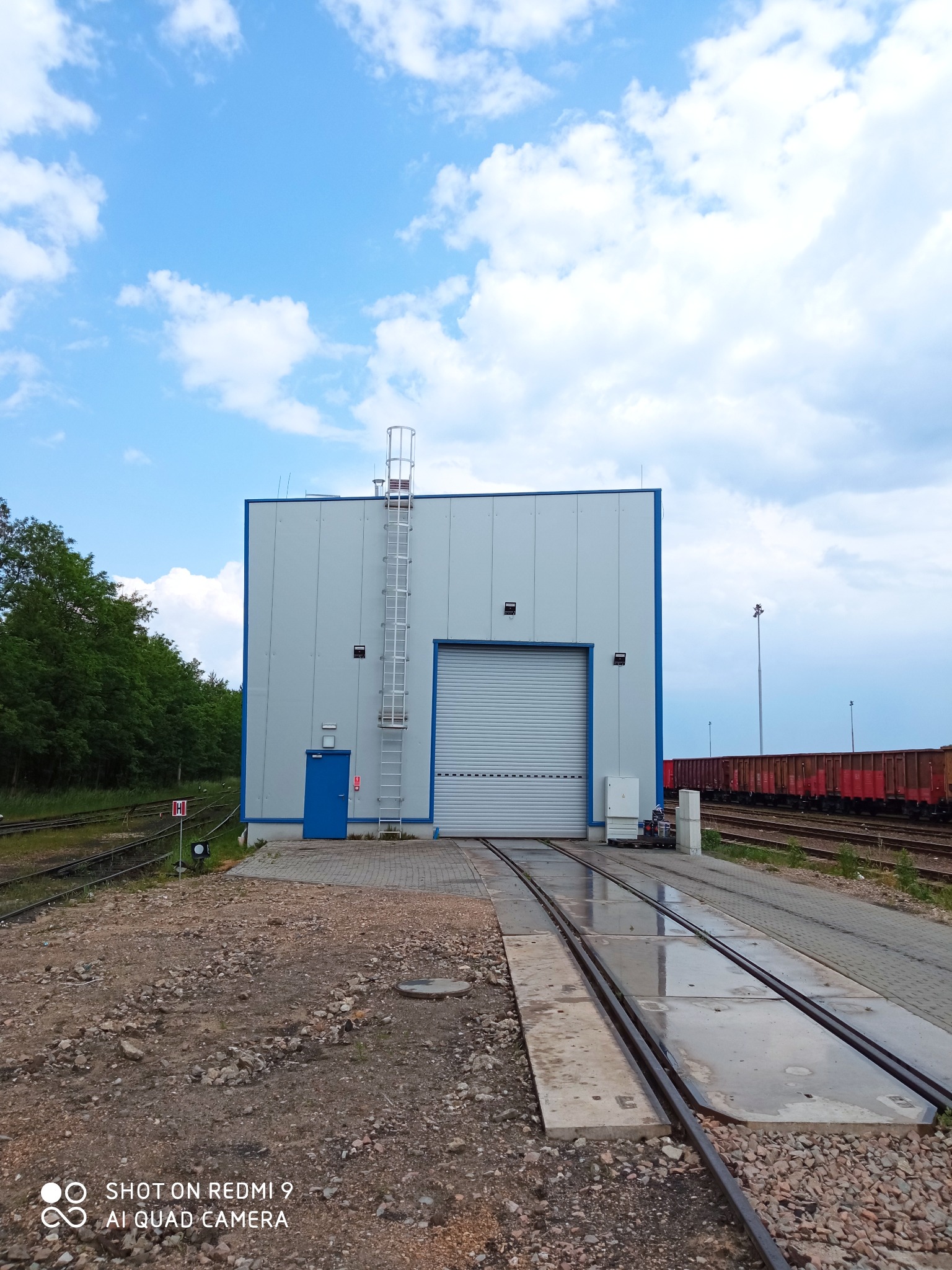 Nowoczesny budynek hali przemysłowej z bramą rolowaną i drabiną na zewnątrz, usytuowany na terenie kolejowym z widocznymi wagonami towarowymi w tle.