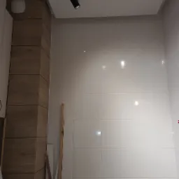 Ściana wyłożona jasnymi, błyszczącymi płytkami ceramicznymi, oświetlona reflektorem sufitowym. Widoczny fragment drewnopodobnej zabudowy i skrzynka z narzędziami w dolnej części kadru.