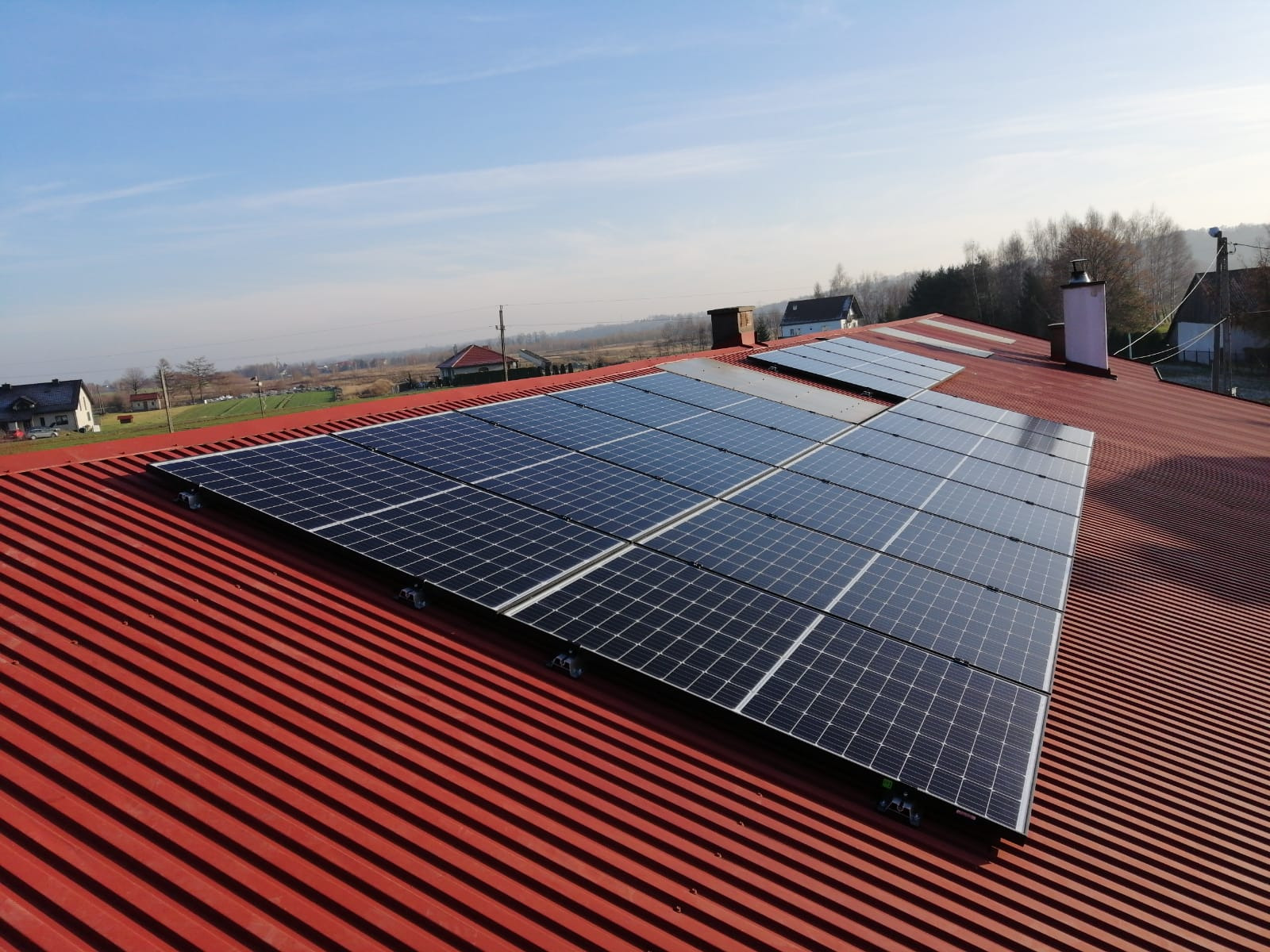 Instalacja fotowoltaiczna na dachu z blachy trapezowej w kolorze czerwonym. Widoczne panele BAUER 320Wp oraz fragment falownika Fronius. W tle zabudowa jednorodzinna i błękitne niebo.