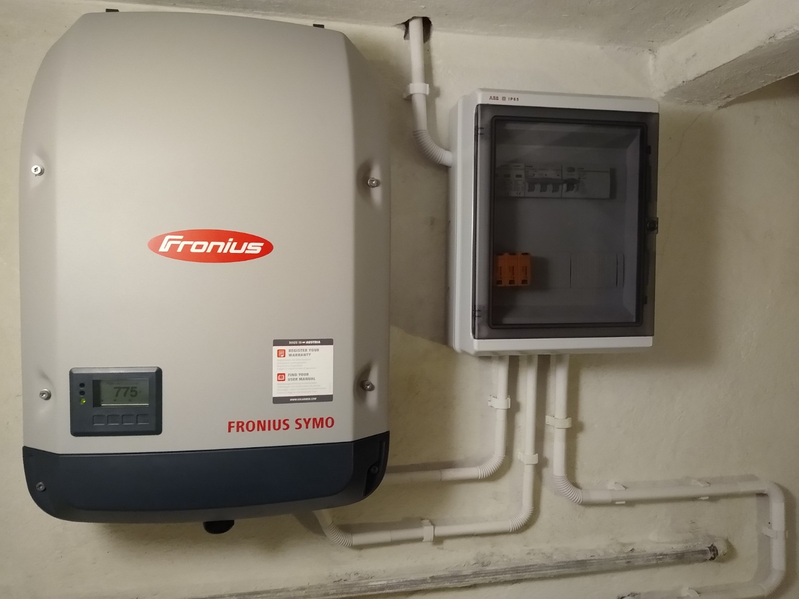 Szary falownik Fronius Symo z wyświetlaczem wskazującym 775, zamontowany obok skrzynki elektrycznej z widocznymi bezpiecznikami, białe rurki instalacyjne prowadzone wzdłuż ściany.