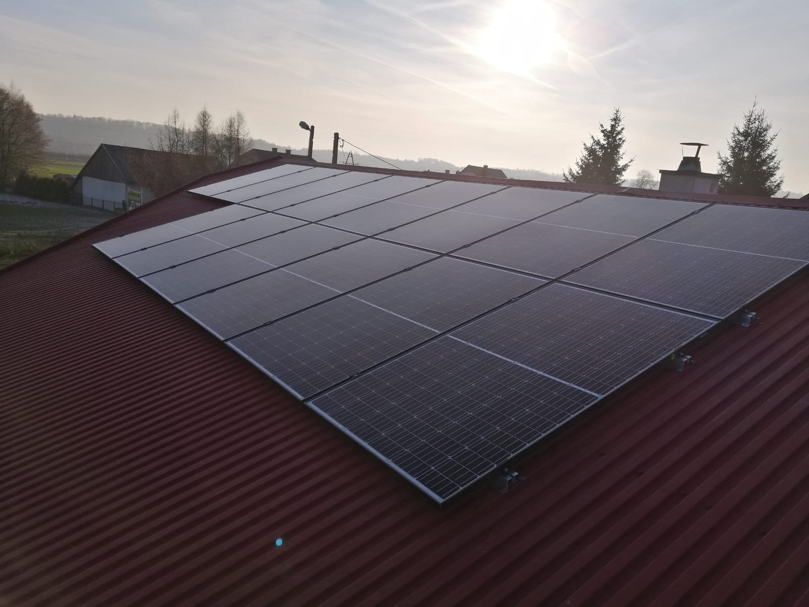 Instalacja fotowoltaiczna na dachu z blachy trapezowej w promieniach zachodzącego słońca. Widoczne panele BAUER ułożone rzędami, z tyłu zabudowania gospodarcze i drzewa.