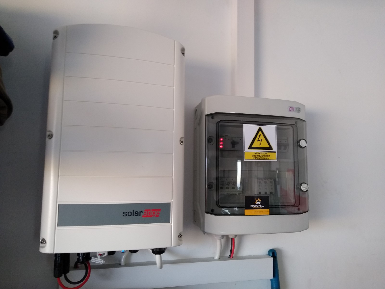 Biały falownik SolarEdge zamontowany na ścianie obok skrzynki elektrycznej z przezroczystymi drzwiczkami i ostrzeżeniem o wysokim napięciu fotowoltaicznym.