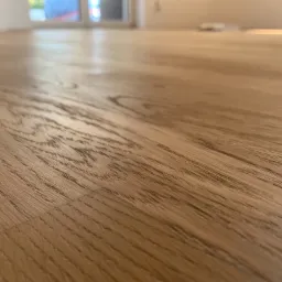 Jasnobrązowa podłoga z wyraźnym rysunkiem słojów drewna, ułożona w nowym, jasnym pomieszczeniu z dużym oknem w tle.